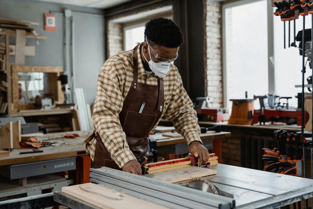Artisan wearing an apron and mask, precisely measuring and cutting wood in a workshop setting.